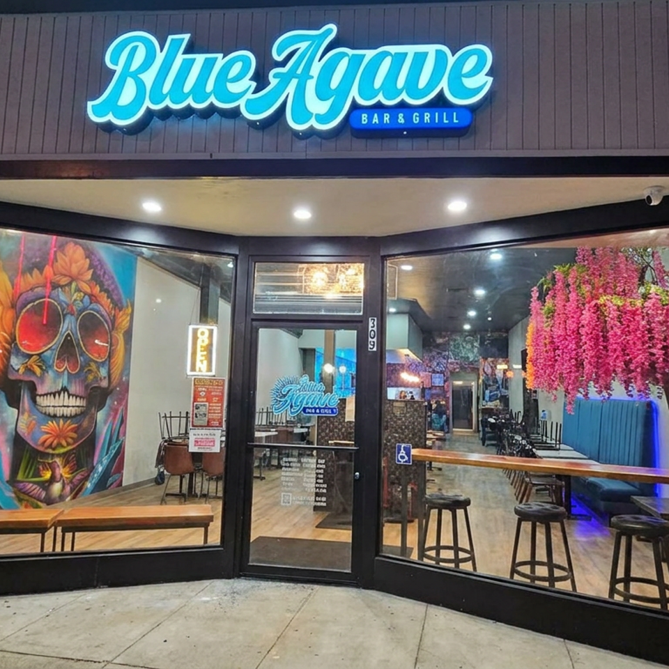 Blue Agave Bar & Grill storefront with colorful skull mural, pink flowering tree, and modern bar seating visible through glass windows.