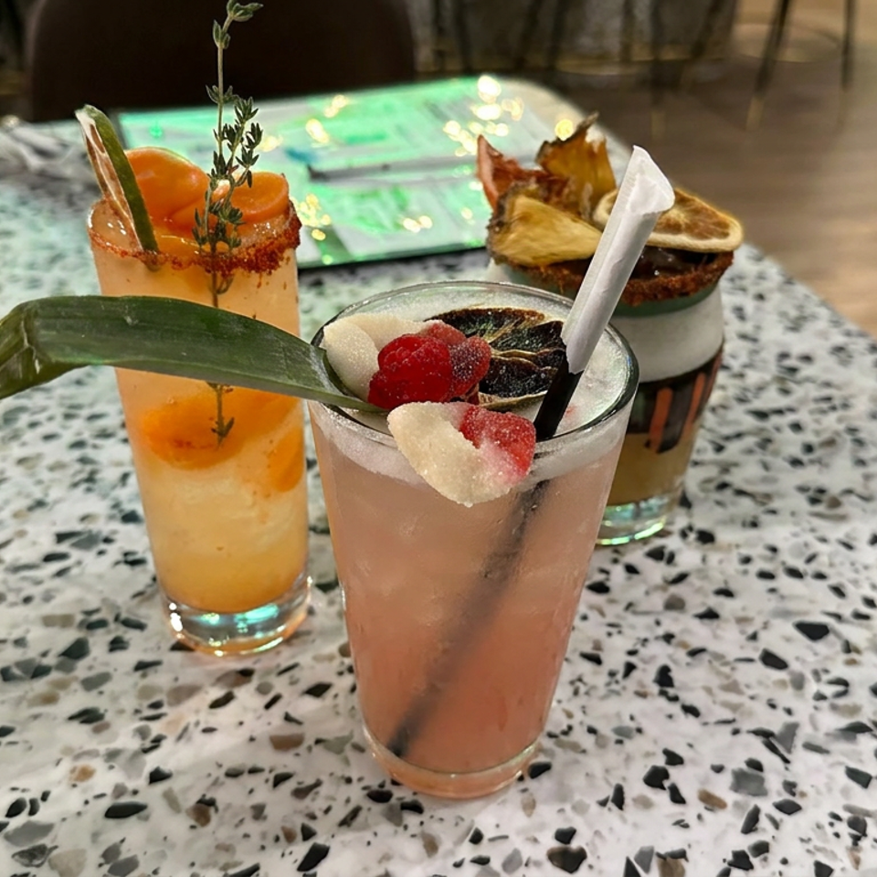 Three colorful cocktails garnished with fruit, herbs, and citrus sit on a terrazzo restaurant table.