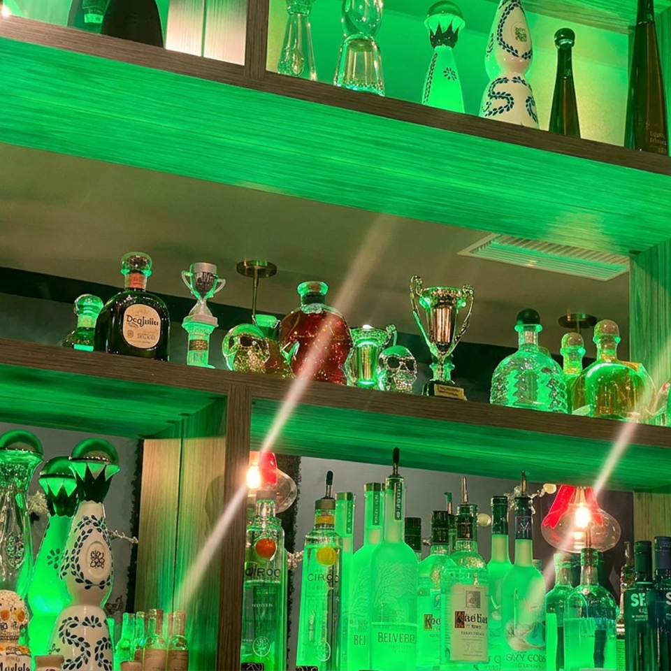 Green-lit restaurant bar shelves displaying liquor bottles, decorative glassware, trophies, and a basketball game on television.